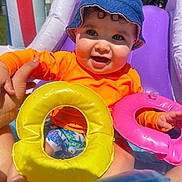 Zayden is registered to the contest to win money with this photo: baby, child, blue_hat, orange_shirt, inflatable_ring, yellow_ring, pink_ring, pool, water, outdoor, summer, sunny, happy, smiling, cute, playing, hand, sitting, curly_hair, fun