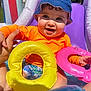 baby, child, blue_hat, orange_shirt, inflatable_ring, yellow_ring, pink_ring, pool, water, outdoor, summer, sunny, happy, smiling, cute, playing, hand, sitting, curly_hair, fun