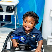 Dainch is registered to the contest to win money with this photo: child, toddler, smiling, toy_car, blue_shirt, ribbon, indoor, happy, face, hands, steering_wheel, short_hair, cute, play, person, seat, clothing, background, baby, portrait