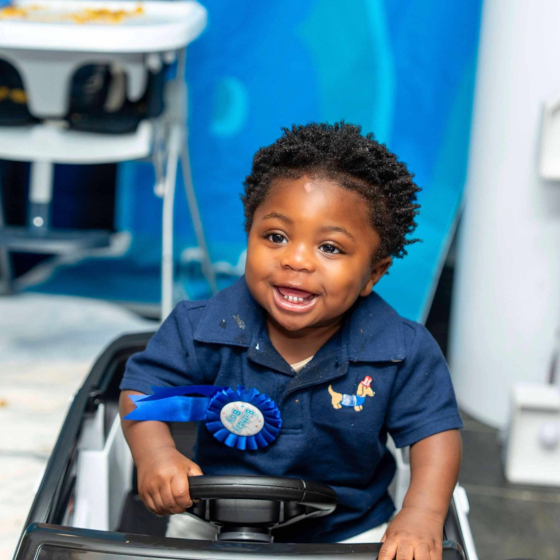 Dainch is registered to the contest to win money with this photo: baby, background, blue_shirt, child, clothing, cute, face, hands, happy, indoor, person, play, portrait, ribbon, seat, short_hair, smiling, steering_wheel, toddler, toy_car