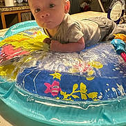 Blake is registered to the contest to win money with this photo: baby, infant, inflatable_ring, colorful, curious, indoor, kitchen, tile_floor, plastic_bottle, clothing, baby_pose, face, child, young, playtime, expression, toy, home, puzzle, floor