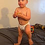 barefoot, bed, blanket, candid, casual, child, diaper, furniture, home, indoor, neutral_expression, person, plain_wall, shadow, skin, small_object, standing, toddler, toy, young_child