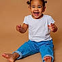 child, toddler, smiling, laughing, sitting, white_tshirt, blue_jeans, barefoot, studio_portrait, beige_background, joyful, expressive_face, hair_buns, earrings, teeth_visible, playful, candid, full_body, cute, portrait