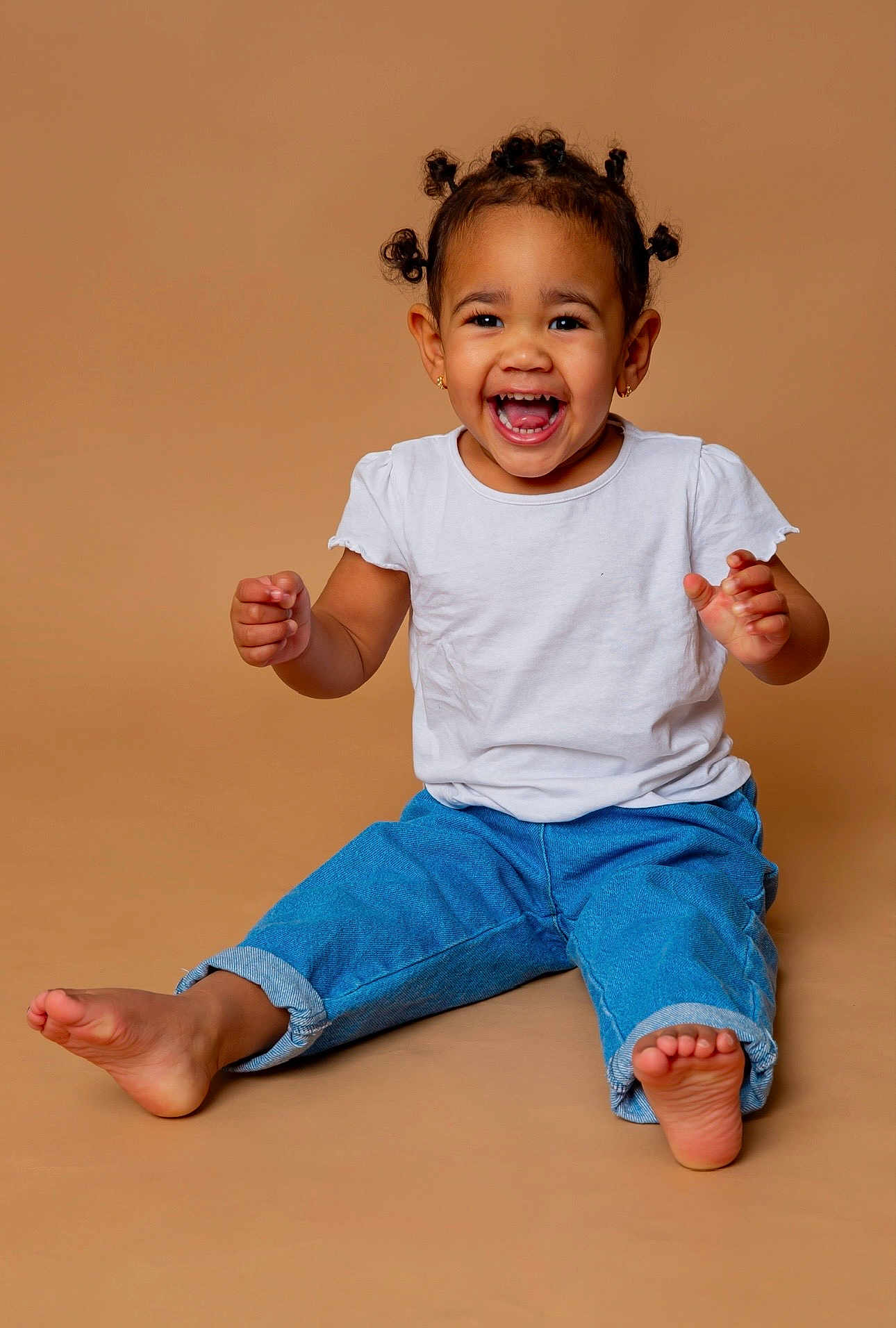 Nïna participe au concours pour gagner de l'argent avec cette photo : child, toddler, smiling, laughing, sitting, white_tshirt, blue_jeans, barefoot, studio_portrait, beige_background, joyful, expressive_face, hair_buns, earrings, teeth_visible, playful, candid, full_body, cute, portrait