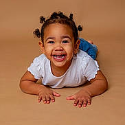 Nïna a rejoint le concours — aidez-le/la à gagner de superbes lots ! child, toddler, smiling, happy, portrait, studio_shot, beige_background, white_shirt, jeans, lying_down, hands, eyes, teeth, curly_hair, hair_buns, earrings, cute, joyful, isolated_background, playful