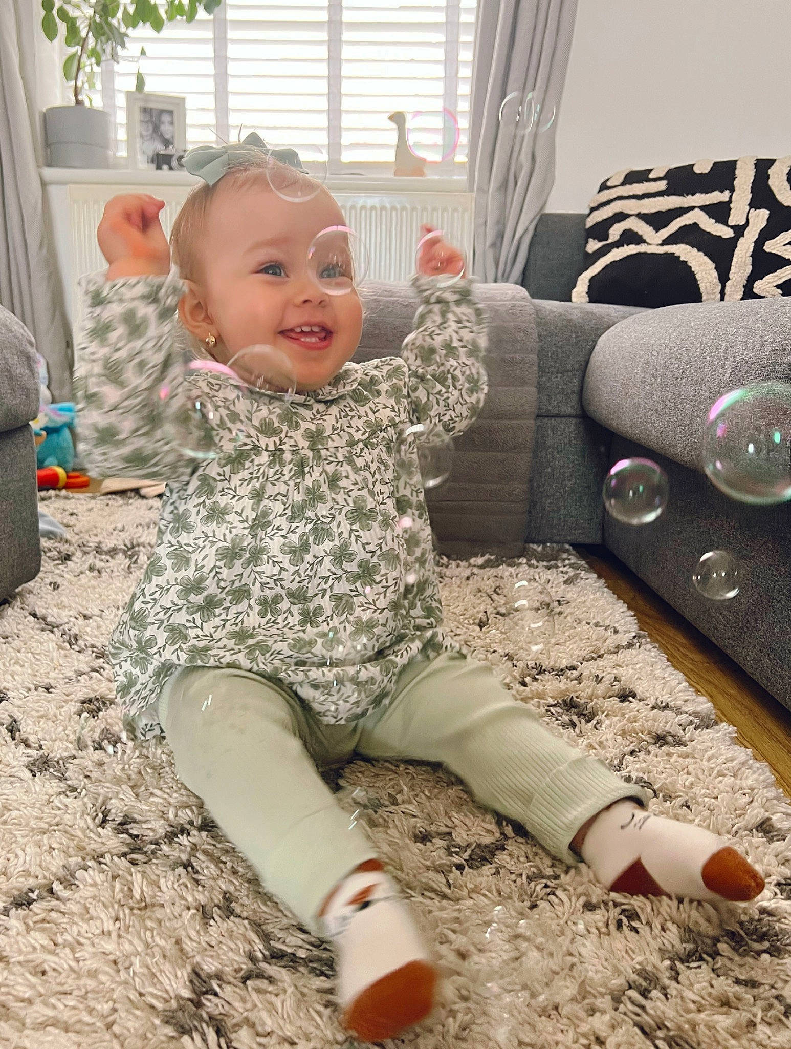 Lara is registered to the contest to win money with this photo: baby, baby_toddler_clothing, chair, child, comfort, couch, floor, flooring, fun, happy, head, human_leg, knee, pattern, person, sitting, smile, thigh, toddler, window_blind