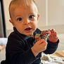 émile a rejoint le concours — aidez-le/la à gagner de superbes lots ! baby, black_shirt, blue_eyes, casual_clothing, child, close_up, colorful_cloth, curious, expression, face, hands, holding, indoor, innocence, pants, portrait, sitting, soft_background, toddler, young_child
