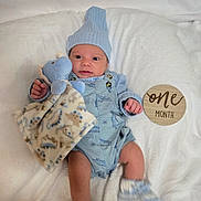 Aaron is registered to the contest to win money with this photo: baby, infant, blue_clothing, knit_hat, plush_toy, soft_blanket, wooden_sign, one_month, newborn, cute, lying_down, baby_booties, child, portrait, indoor, smiling, baby_girl_or_boy, milestone, cozy, resting
