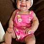 baby, smiling, headband, bow, romper, pink_clothing, daisy_pattern, sitting, barefoot, chubby_arms, chubby_legs, brown_blanket, infant, happy, indoor, cute, portrait, child, one_person, closeup
