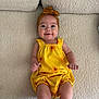 baby, infant, smiling, yellow_clothing, headband, sunflower_pattern, couch, textured_fabric, happy, cute, portrait, child, indoors, sitting, comfort, skin, human, one_person, young_child, playful