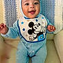 baby, child, smiling, blue_onesie, mickey_mouse, bib, knitted_blanket, chair, happy, infant, portrait, seated, cute, indoors, cozy, face, hands, clothing, babywear, soft_texture