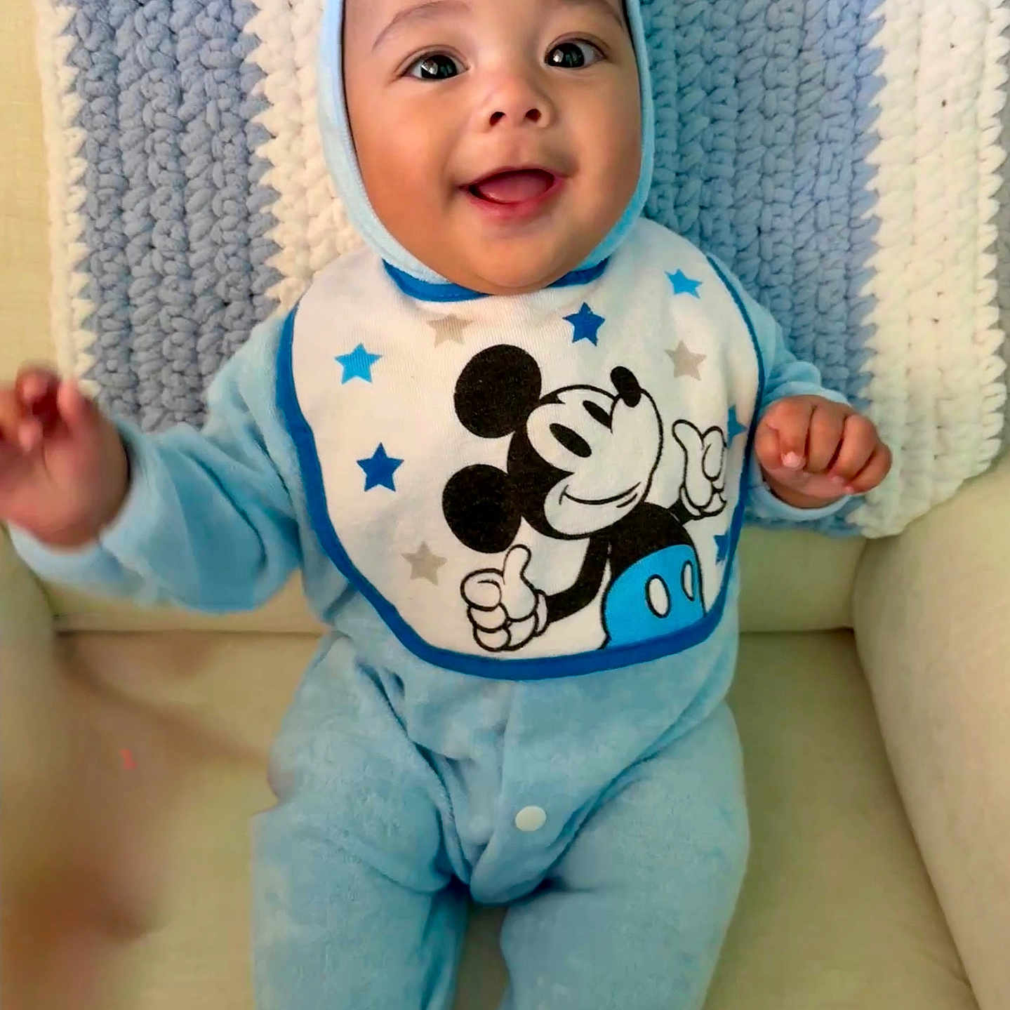 Teodoro is registered to the contest to win money with this photo: baby, babywear, bib, blue_onesie, chair, child, clothing, cozy, cute, face, hands, happy, indoors, infant, knitted_blanket, mickey_mouse, portrait, seated, smiling, soft_texture