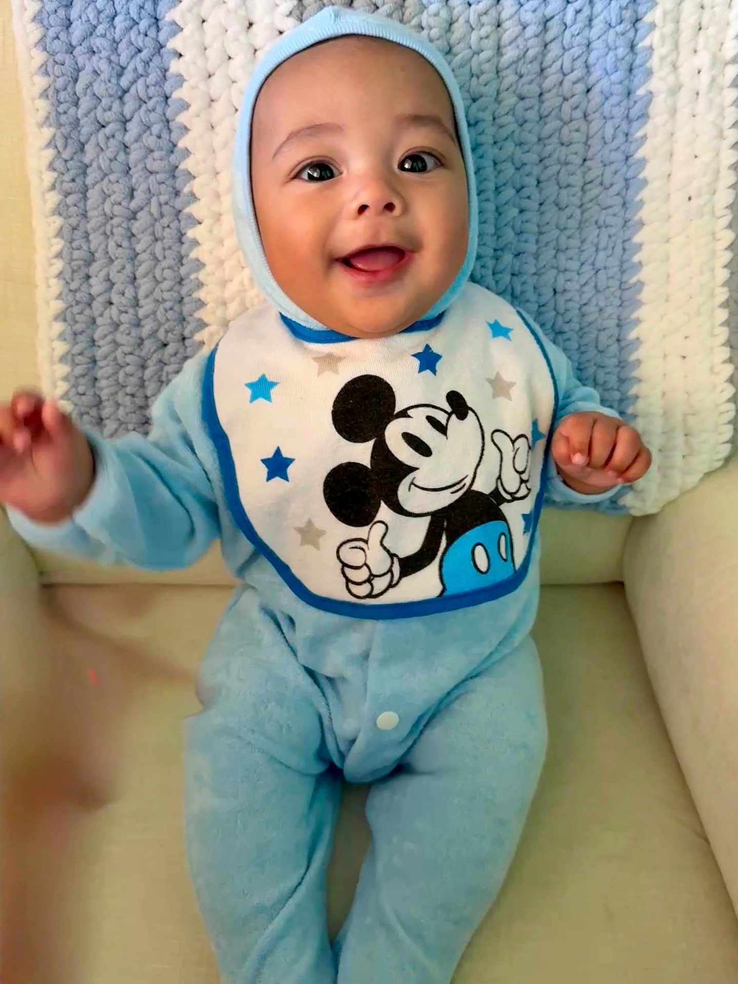 Teodoro is registered to the contest to win money with this photo: baby, child, smiling, blue_onesie, mickey_mouse, bib, knitted_blanket, chair, happy, infant, portrait, seated, cute, indoors, cozy, face, hands, clothing, babywear, soft_texture