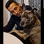 man, dog, indoor, cuddle, table, curtains, brindle, pet, companion, affection, seated, closeup, portrait, bond, warm_lighting, casual_clothing, domestic, animal, friendship, relaxed