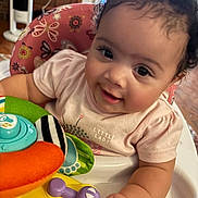 Eliza is registered to the contest to win money with this photo: baby, child, smiling, curly_hair, high_chair, toy, indoor, cute, person, infant, playful, colorful, pink_shirt, face, happy, young_child, closeup, skin, hand, expression