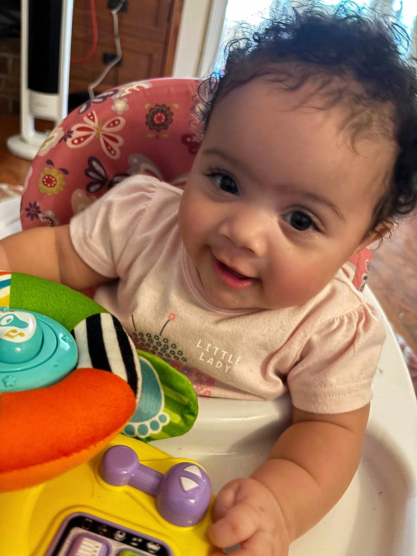 Eliza is registered to the contest to win money with this photo: baby, child, smiling, curly_hair, high_chair, toy, indoor, cute, person, infant, playful, colorful, pink_shirt, face, happy, young_child, closeup, skin, hand, expression