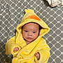 Luna joined the competition — help win amazing prizes! baby, child, yellow_robe, duck_costume, hood, bedspread, patterned_bed, infant, cute, fuzzy_clothing, indoor, portrait, face, hands, lying_down, soft_texture, sleepwear, cozy, small_fingers, expression