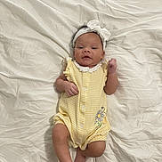 Luna joined the competition — help win amazing prizes! baby, infant, child, headband, white_bow, yellow_romper, striped_clothing, daisy_embroidery, lying_down, bed_sheet, white_sheet, curly_hair, cute, portrait, skin, foot, hand, indoors, young_child, smiling