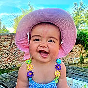 Lesley is registered to the contest to win money with this photo: baby, child, smiling, hat, pink_hat, outdoor, wooden_table, stone_wall, greenery, sunlight, blue_sky, colorful_clothing, flower_straps, happy, portrait, sitting, cute, daylight, nature, person