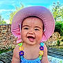 baby, child, smiling, hat, pink_hat, outdoor, wooden_table, stone_wall, greenery, sunlight, blue_sky, colorful_clothing, flower_straps, happy, portrait, sitting, cute, daylight, nature, person