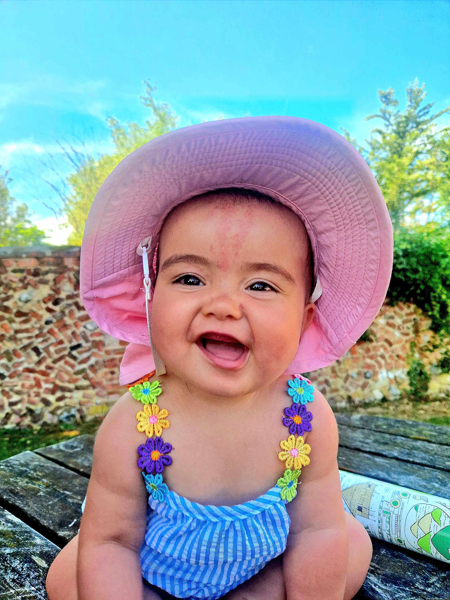 Lesley is registered to the contest to win money with this photo: baby, child, smiling, hat, pink_hat, outdoor, wooden_table, stone_wall, greenery, sunlight, blue_sky, colorful_clothing, flower_straps, happy, portrait, sitting, cute, daylight, nature, person