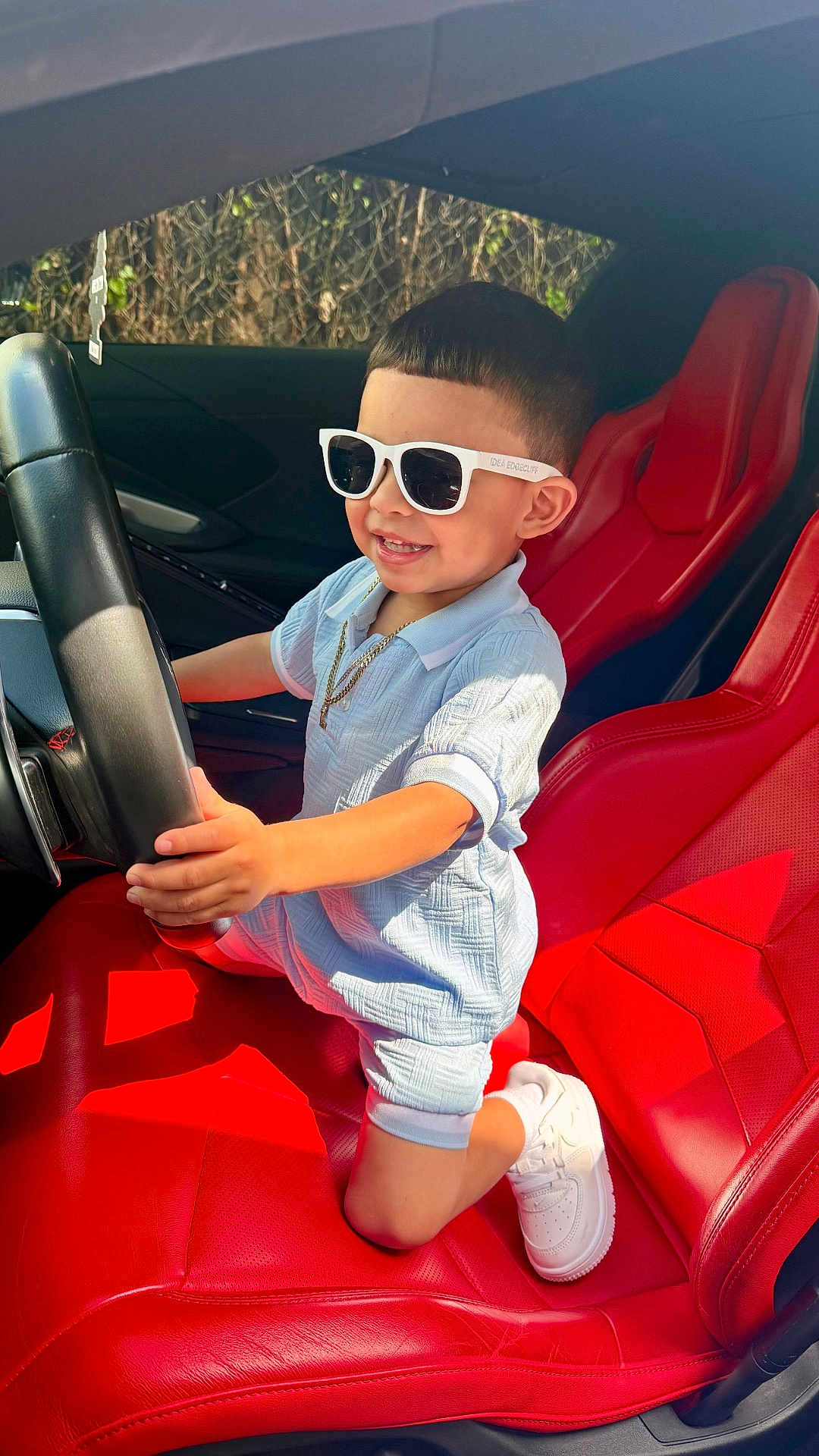 Liam is registered to the contest to win money with this photo: child, boy, sunglasses, car, steering_wheel, red_leather_seat, white_shoes, smiling, short_hair, casual_clothing, sunlight, interior, vehicle, seat, gold_chain, happy, portrait, daylight, cute, playful