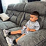 Liam joined the competition — help win amazing prizes! child, boy, video_game_controller, gray_couch, sneakers, shorts, tshirt, gold_chain, haircut, curtains, pillow, carpet, indoors, playing, focused, seated, furniture, person, living_room, casual