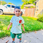 Liam is registered to the contest to win money with this photo: toddler, child, boy, outdoor, sidewalk, grass, sneakers, shorts, tshirt, sunlight, smile, sunglasses, chain, bracelet, vehicle, fence, tree, daytime, playful, casual