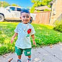 toddler, child, boy, outdoor, sidewalk, grass, sneakers, shorts, tshirt, sunlight, smile, sunglasses, chain, bracelet, vehicle, fence, tree, daytime, playful, casual