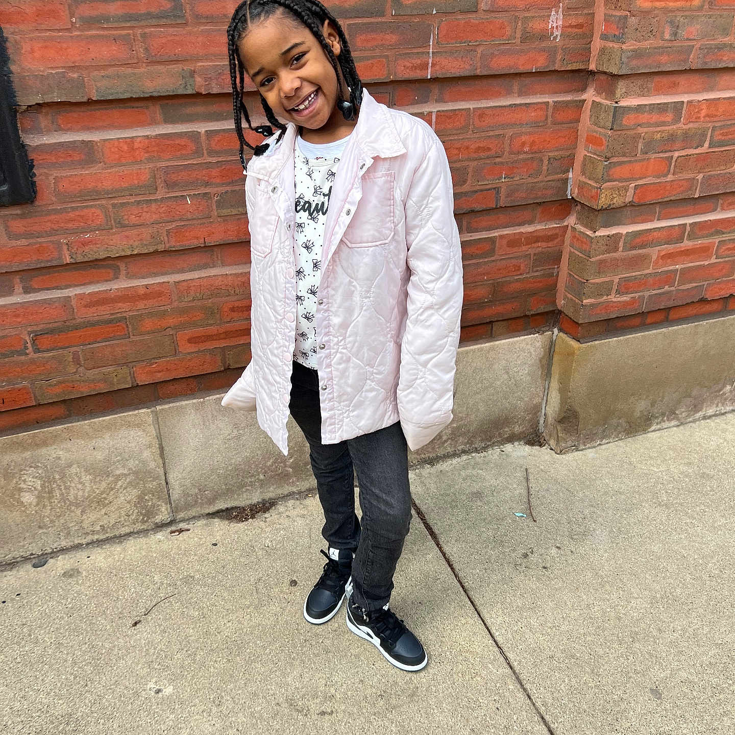 Tamiera is registered to the contest to win money with this photo: braids, brick_wall, casual_clothing, child, concrete, cute, daylight, fashion, happy, jacket, outdoor, person, playful, portrait, sidewalk, smile, sneakers, standing, urban, young