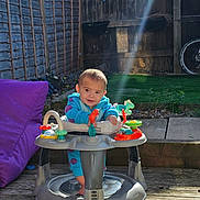 Theo-John joined the competition — help win amazing prizes! baby, child, walker, outdoor, sunlight, wooden_deck, toy, activity_center, purple_cushion, grass, fence, garden, footwear, clothing, standing, play, daylight, cute, infant, person