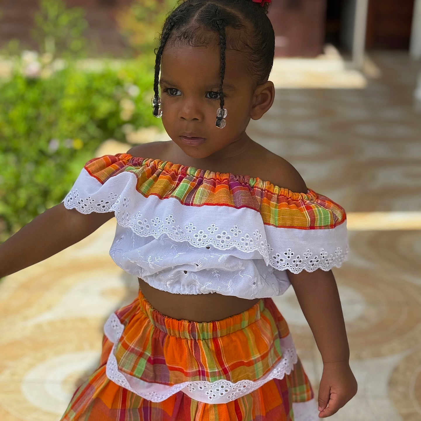 Naëlys a rejoint le concours — aidez-le/la à gagner de superbes lots ! child, girl, plaid, colorful_clothing, lace, braids, beads, outdoor, garden, patterned_floor, standing, portrait, fashion, young_child, person, sunlight, hair_bun, serious_expression, summer_outfit, toddler