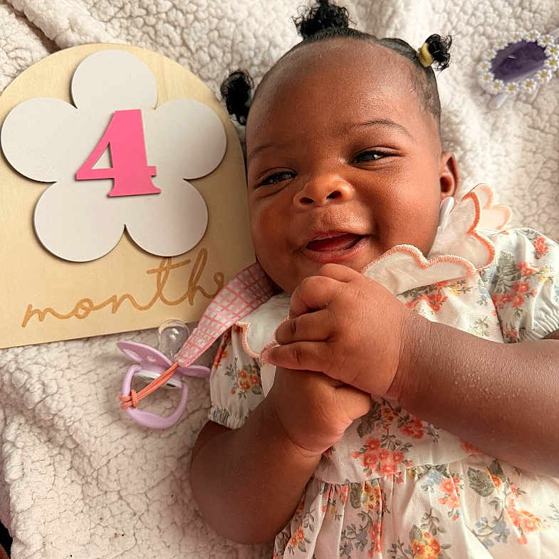 Naioir is registered to the contest to win money with this photo: baby, infant, smiling, girl, child, floral_dress, blanket, hair_puffs, cute, happy, lying_down, indoors, sign, four_months, portrait, skin, hands, close_up, young_child, adorable