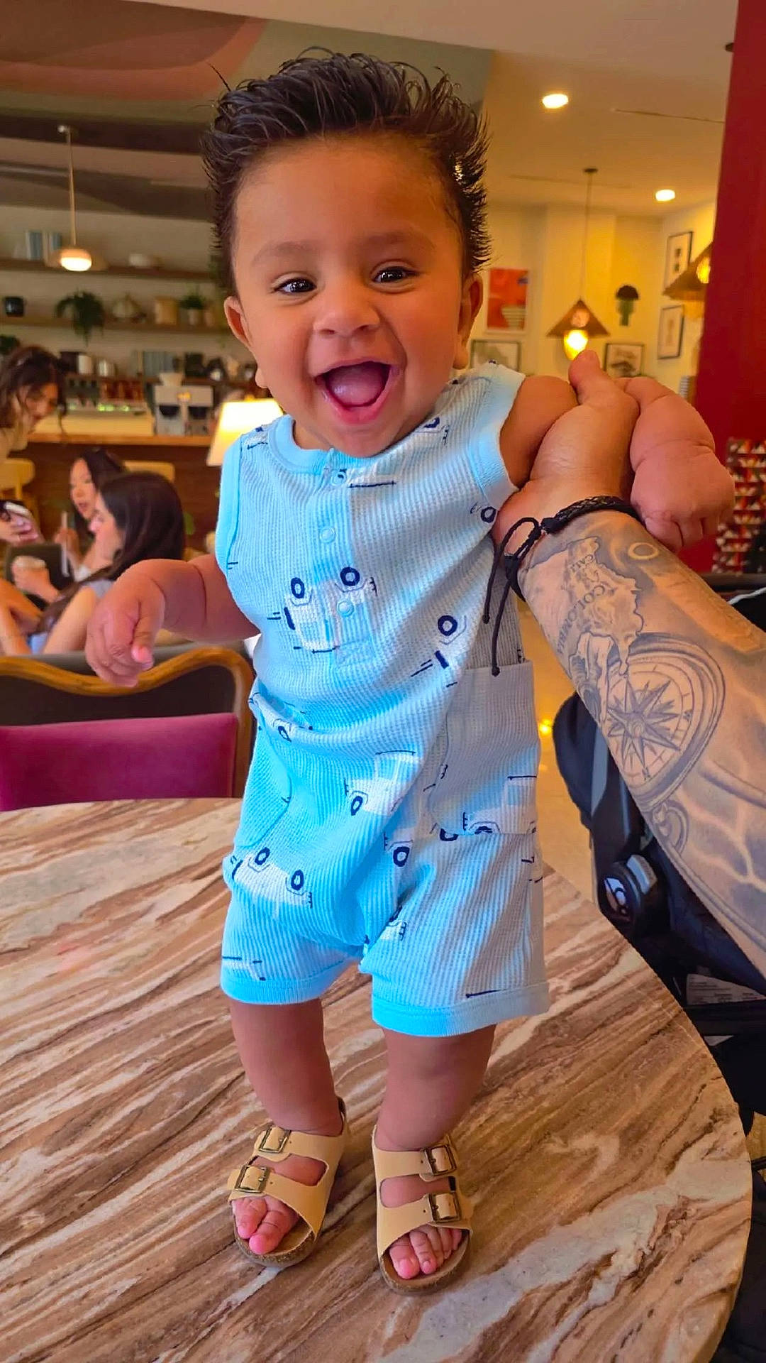 Ricky is registered to the contest to win money with this photo: baby, background, blue_romper, cafe, child, cute, expression, footwear, hair, hand, happy, indoor, marble_table, people, person, portrait, sandals, smiling, standing, tattoo