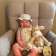 Henry is registered to the contest to win money with this photo: baby, child, smiling, hat, overalls, stuffed_animal, giraffe_toy, armchair, plush_furniture, milestone_sign, wooden_sign, indoor, cute, happy, infant, clothing, person, portrait, sitting, home