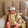 baby, child, smiling, hat, overalls, stuffed_animal, giraffe_toy, armchair, plush_furniture, milestone_sign, wooden_sign, indoor, cute, happy, infant, clothing, person, portrait, sitting, home