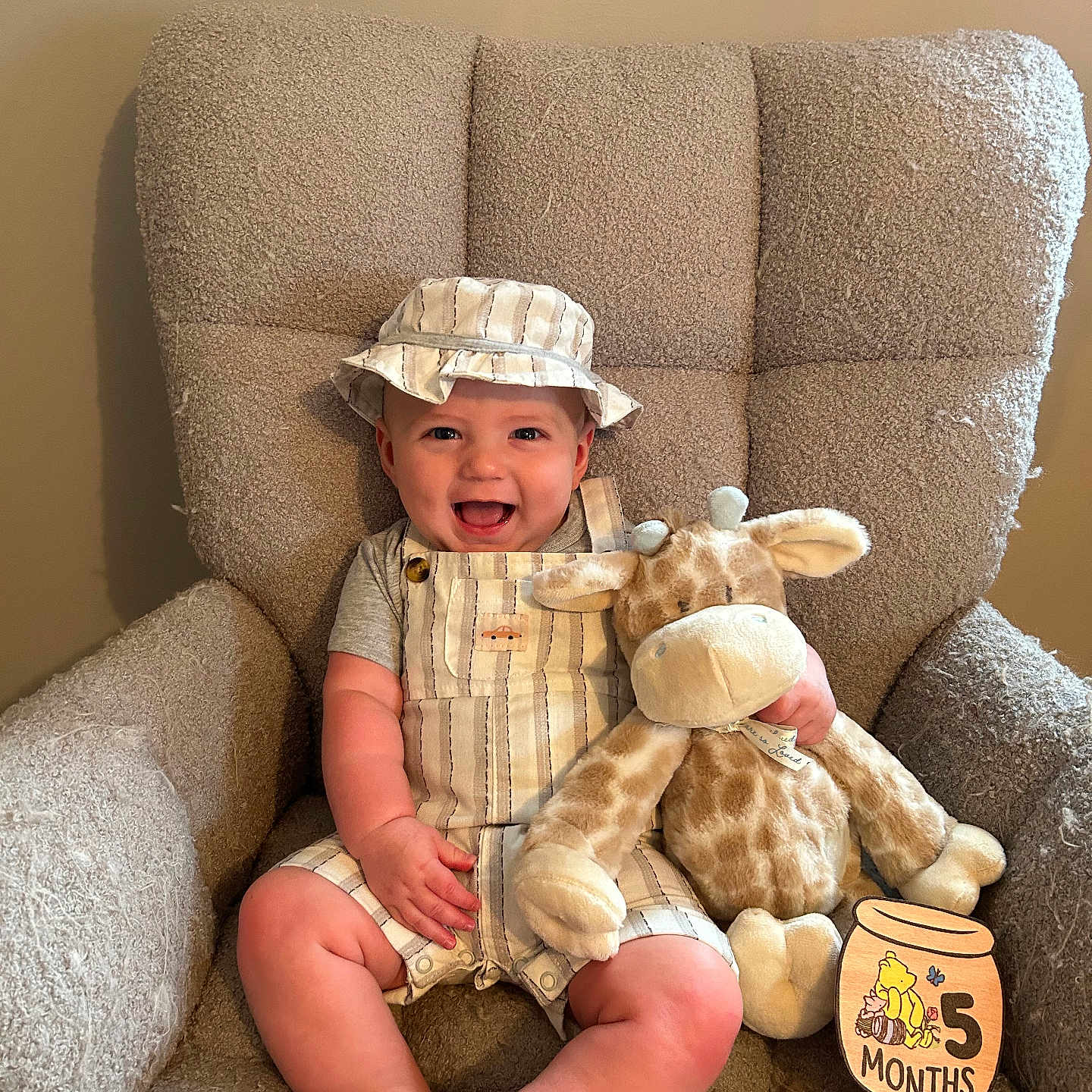 Henry is registered to the contest to win money with this photo: baby, child, smiling, hat, overalls, stuffed_animal, giraffe_toy, armchair, plush_furniture, milestone_sign, wooden_sign, indoor, cute, happy, infant, clothing, person, portrait, sitting, home