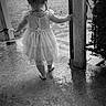 toddler, child, barefoot, dress, wet_floor, porch, black_and_white, outdoor, back_view, post, rope, grass, concrete, small_person, young_child, single_child, nature, daylight, curly_hair, walking