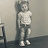 toddler, child, girl, standing, shirt, hair_bows, jeans, sneakers, chair, wall, floor, shadow, indoor, monochrome, vintage, smile, positive_message, casual_clothing, portrait, young_child
