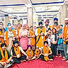 adults, beard, carpet, celebration, children, community, culture, diverse_ages, group, headwear, indoor, orange_scarf, people, portrait, posing, religious_gathering, sikh, smiling, traditional_clothing, turban