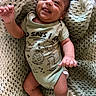 baby, infant, smiling, tongue_out, onesie, dinosaur_print, knitted_blanket, cozy, cute, happy, chubby_arm, lying_down, indoors, child, person, skin, head, hand, foot, red_bracelet