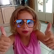 Océane a rejoint le concours — aidez-le/la à gagner de superbes lots ! child, glasses, thumbs_up, pink_shirt, indoor, kitchen, tile_floor, blonde_hair, smiling, person, casual, home, door, clothes, reflection, natural_light, portrait, young_child, positive_gesture, fashion