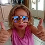 child, glasses, thumbs_up, pink_shirt, indoor, kitchen, tile_floor, blonde_hair, smiling, person, casual, home, door, clothes, reflection, natural_light, portrait, young_child, positive_gesture, fashion