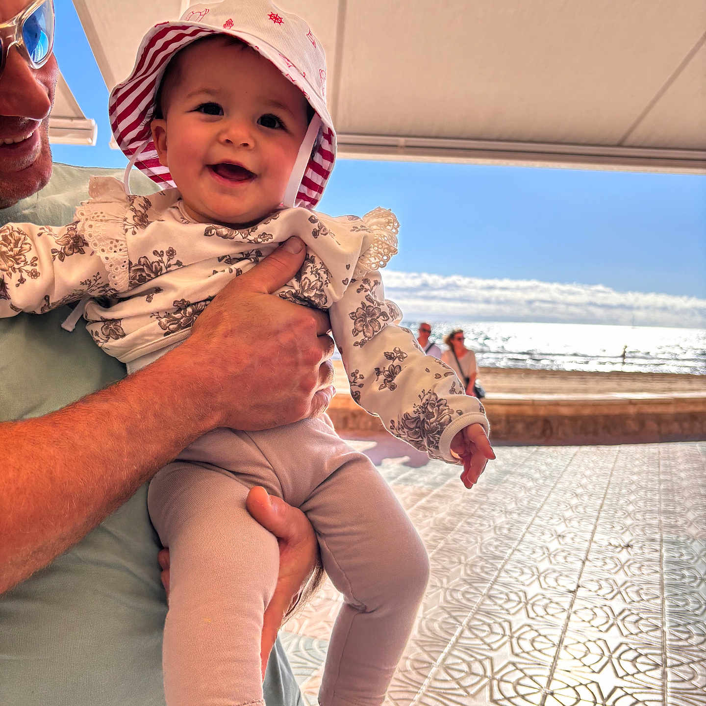 Alina participe au concours pour gagner de l'argent avec cette photo : adult_hand, baby, beach, canopy, child, clothing, daylight, happy, hat, holding, ocean, outdoor, patterned_floor, person, portrait, shadow, smiling, sun_hat, sunlight, tile_floor