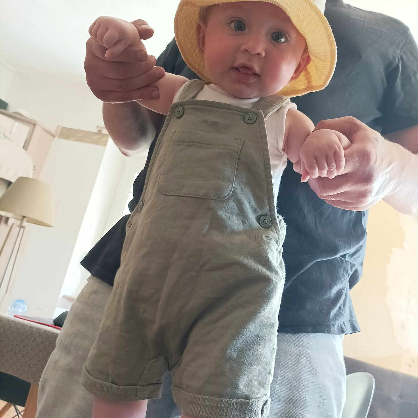 Charly participe au concours pour gagner de l'argent avec cette photo : baby, child, person, adult, hat, bucket_hat, overalls, green_overalls, indoor, furniture, lamp, smiling, holding_hands, standing, learning_to_walk, home, casual_clothing, beard, man, happy