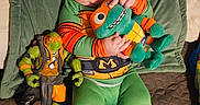Roman is registered to the contest to win money with this photo: baby, child, smiling, plush_toy, action_figure, green_clothing, beanie, toddler, indoor, quilted_bedspread, toy, cute, happy, playful, person, cushion, footwear, orange, green, portrait