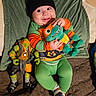 baby, child, smiling, plush_toy, action_figure, green_clothing, beanie, toddler, indoor, quilted_bedspread, toy, cute, happy, playful, person, cushion, footwear, orange, green, portrait