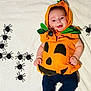 baby, pumpkin_costume, halloween, toy_spiders, orange, green_leaves, white_blanket, smiling, cute, infant, child, costume, holiday, playful, lying_down, blue_pants, festive, decoration, indoors, portrait
