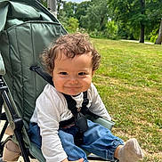 Nael participe au concours pour gagner de l'argent avec cette photo : toddler, child, stroller, outdoor, grass, trees, curly_hair, smile, jeans, socks, white_shirt, playful, nature, park, baby, seatbelt, daylight, person, happy, cute