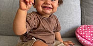 Nael participe au concours pour gagner de l'argent avec cette photo : toddler, child, smiling, sitting, curly_hair, brown_shirt, beige_shorts, white_shoes, couch, indoor, home, happy, face, hand, footwear, staircase, pink_cushion, polka_dot, person, portrait